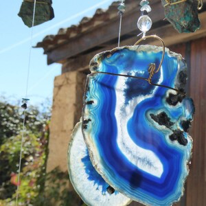 May include: A close-up of a blue and white agate slice wind chime. The agate slices are suspended by wire and adorned with beads. The agate has a striking blue color with white and black accents. Other hanging stones are visible in the background.