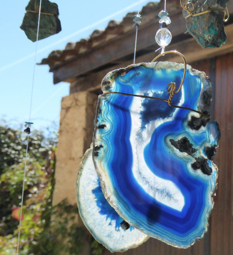May include: A close-up of a blue and white agate slice wind chime. The agate slices are suspended by wire and adorned with beads. The agate has a striking blue color with white and black accents. Other hanging stones are visible in the background.