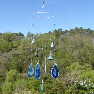 May include: A handcrafted wind chime featuring blue and white agate slices and various blue and turquoise stones suspended from a circular metal frame. The wind chime hangs outdoors against a backdrop of green trees and a blue sky.