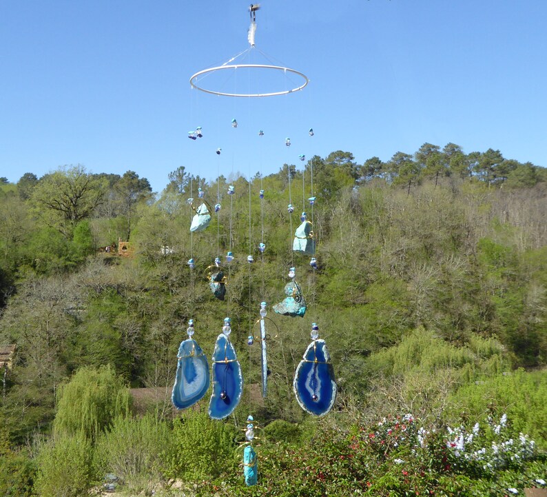 May include: A handcrafted wind chime featuring blue and white agate slices and various blue and turquoise stones suspended from a circular metal frame. The wind chime hangs outdoors against a backdrop of green trees and a blue sky.