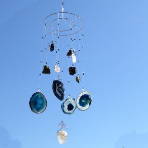 May include: A hanging mobile with a silver metal frame and multiple strands of various sized, polished gemstones. The gemstones include blue agate slices, clear quartz, and dark green and black stones, all suspended against a clear blue sky.