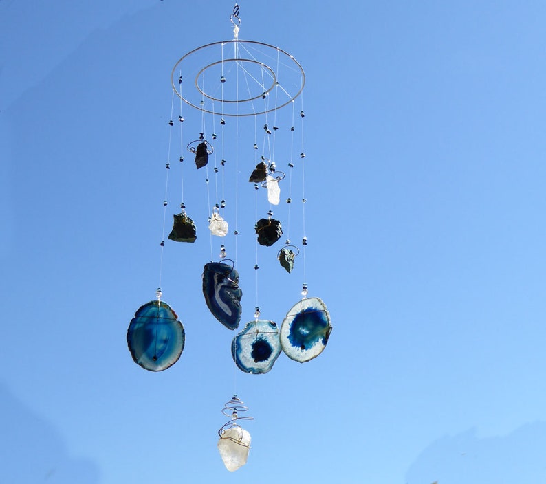 May include: A hanging mobile with a silver metal frame and multiple strands of various sized, polished gemstones. The gemstones include blue agate slices, clear quartz, and dark green and black stones, all suspended against a clear blue sky.