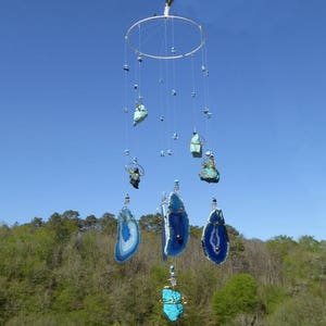 May include: A handcrafted wind chime featuring a circular metal frame with multiple strands of varying lengths. Each strand holds a combination of blue and turquoise-colored stones and polished agate slices, creating a decorative outdoor accent.