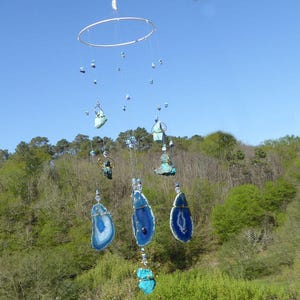 May include: A handcrafted mobile featuring blue and white agate slices, turquoise stones, and small blue beads suspended from a silver ring. The mobile hangs against a backdrop of a bright blue sky and lush green trees.