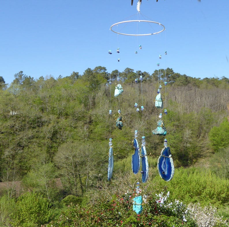 May include: A handcrafted wind chime with a circular top and multiple strands of decorative elements. The wind chime features blue, turquoise, and clear accents, with a natural outdoor background of trees and a blue sky.