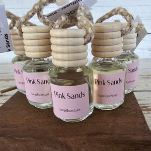 May include: Six clear glass bottles with wooden stoppers and pink labels that read "Pink Sands" and "NanasEssentials". The bottles are hanging from a rope and are on a wooden surface.