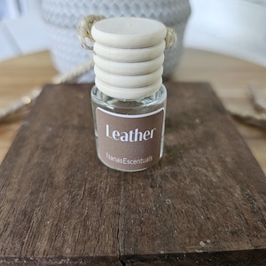 May include: A small, clear glass bottle with a wooden cap and a brown label that reads "Leather" and "NanasEscentuals". The bottle sits on a dark brown wooden surface. The cap is light-colored and has a rope attached.