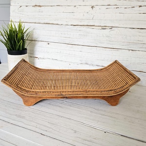 May include: A long, rectangular bamboo tray with a curved design. The tray is made of woven bamboo and has a light brown color. It sits on a wooden base with four small feet. The tray is approximately 28 inches long.
