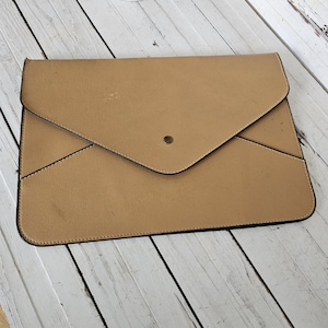 May include: Tan envelope-style clutch with a flap closure and a single brass-colored button. The clutch has black trim and a stitched border. The surface shows minor wear and tear.