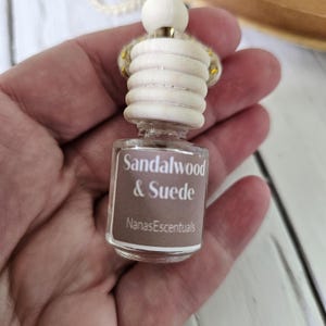 May include: A small, clear glass bottle with a wooden top and a label that reads "Sandalwood & Suede" and "NanasEscentuals". The bottle is held in a hand and is designed to be an air freshener or fragrance diffuser.