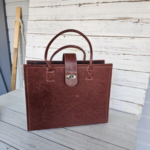 May include: A brown leather briefcase with a handle and a silver clasp. The briefcase has a rectangular shape with white stitching around the edges. The handle is arched and made of the same brown leather.
