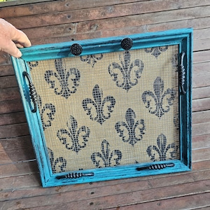 May include: A rectangular, turquoise-painted screen with a burlap-like interior featuring a black fleur-de-lis pattern. The screen has black handles and knobs. The frame is distressed, giving it a rustic appearance.