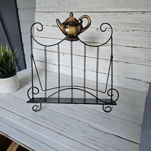 May include: Black metal cookbook stand with a decorative teapot on top. The stand has a curved base and top with chain accents. The teapot is brown and gold. The stand is on a light wood surface.