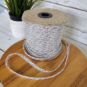 May include: A spool of braided cord in shades of gray and white. The cord is wound around a cardboard core with a black center. A length of the cord is unraveled and lies on a wooden surface. A small potted plant is in the background.