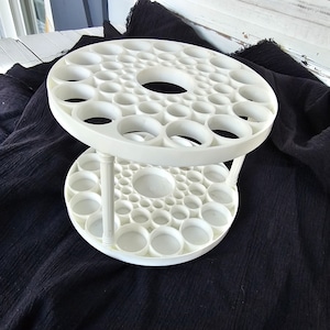 May include: A two-tiered, white, circular display stand with numerous circular openings. The stand is supported by three white posts and is set against a dark background. The top tier has a large central opening.