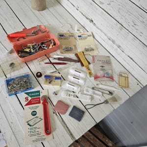 May include: A collection of sewing supplies including a coral plastic case filled with metal presser feet, Dritz snap fastener packages, a yellow measuring tape, Singer bobbins, a red Singer tracing wheel, white plastic attachments, and pins.
