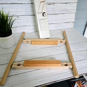May include: A wooden embroidery hoop stand, partially assembled, with two wooden bars and two side supports. The stand is light-colored wood with metal hardware. A box with product information is visible in the background.