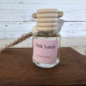 May include: A clear glass bottle with a pink label that says "Pink Sands" and "NanasEssentials". The bottle has a wooden cap with a rope attached to it.