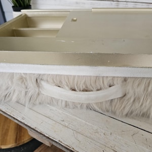 May include: A light beige vanity case with a faux fur exterior and a white handle. The case has a gold-colored interior with compartments and a hinged lid. The case is resting on a distressed white wooden surface.