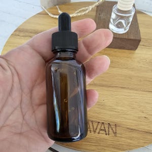 May include: A brown glass dropper bottle with a black dropper top is held in a hand. The bottle is partially filled with a clear liquid. A small glass bottle and a wooden block are in the background.