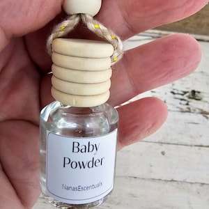 May include: A clear glass bottle with a white label that reads "Baby Powder" and "NanasEscentials". The bottle is hanging from a white wooden bead and a rope with yellow accents.