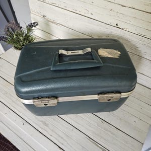 May include: A dark teal, rectangular hard-shell travel case with a textured exterior. The case features a central carrying handle with the brand name Magnalite, two silver-toned metal latches on the front, and a small, worn adhesive label.