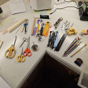 May include: A collection of crafting tools including various scissors, metal sculpting ball styluses, precision knives, tweezers, paintbrushes, sponges, and markers arranged on a surface. The set features yellow, orange, blue, and black handles.