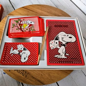 May include: Vintage Snoopy card game set. The set includes a deck of red and white checkered cards, a score pad with Snoopy holding cards, and a gold pen. The cards and score pad have a red polka dot background. The set is in a white box.