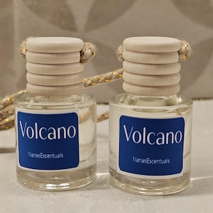 May include: Two small, clear glass bottles with wooden caps and blue labels that read "Volcano" and "NanasEscentuals". The bottles are connected by a gold-colored string.