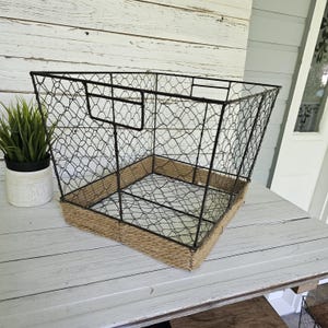 May include: A large, open wire basket with a rectangular base and a trapezoidal shape. The basket is made of black metal wire with a woven rope trim around the base. A small potted plant sits to the left.