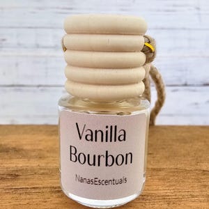 May include: A small glass bottle with a wooden cap and a string hanging from it. The bottle contains a clear liquid and has a label that reads "Vanilla Bourbon" and "NanasEscentials".
