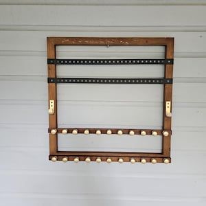 May include: A wooden jewelry organizer with a dark brown frame. It features two black bars with evenly spaced holes and two rows of gold knobs. Two gold hooks are on the sides. The organizer is mounted on a white, horizontal-planked wall.