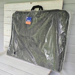 May include: A black mesh art portfolio in a clear plastic sleeve with a black handle. A blue and orange tag is attached to the handle. The portfolio is leaning against a white wooden surface.