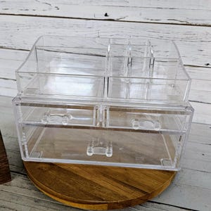 May include: Clear acrylic makeup organizer with multiple compartments and two drawers. The organizer sits on a round, wooden base. The top section has various slots for brushes and bottles, while the drawers offer additional storage.