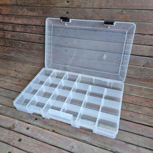 May include: A clear plastic organizer box with a hinged lid and multiple compartments. The box is open, revealing the interior dividers. The lid has two black latches. The box is on a wooden surface.