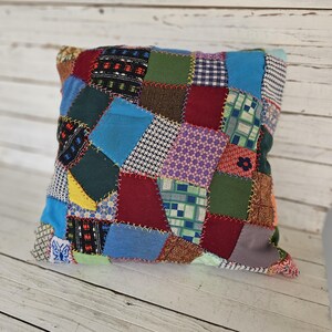 May include: Square patchwork pillow with a variety of fabric patterns and colors. The pillow features a mix of geometric shapes, floral prints, and solid colors, including blue, red, green, and brown. The seams are visible, adding to the handmade aesthetic.