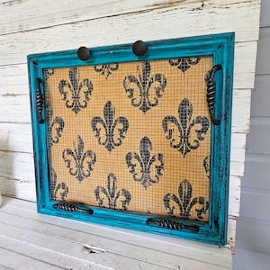 May include: A rectangular decorative screen with a teal frame and black hardware. The screen features a tan mesh background with a black fleur-de-lis pattern. The frame has two black knobs and two twisted metal handles.
