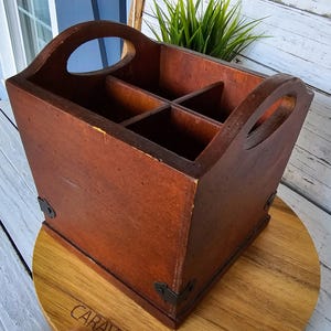 May include: A wooden caddy with four compartments and a handle on each end. The caddy is stained a warm brown color and has metal corner brackets. The caddy is sitting on a wooden surface.