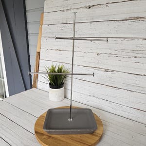 May include: A silver-colored, tiered jewelry display stand with a rectangular base. The stand has two horizontal bars for hanging jewelry and a central vertical support. The base is gray and sits on a wooden round base.