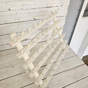 May include: A cream-colored wooden thread rack with multiple rows of pegs. The rack is designed to hold spools of thread, with each peg providing a spot for a spool. The rack is angled and has a vintage aesthetic.