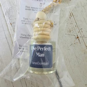May include: A small clear glass bottle with a wooden cap and hanging cord, packaged in a clear plastic bag with an instruction card. The blue label on the bottle features the text The Perfect Man and NanasEscentuals in white lettering.