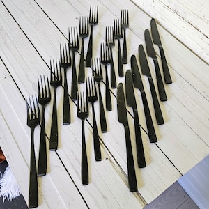 May include: An assortment of black forks and knives arranged on a white wooden surface. The forks have four tines, and the knives have a simple, sleek design. The flatware is made of a dark, matte material, creating a modern aesthetic.