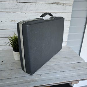 May include: A vintage black briefcase with a textured surface and silver trim. The briefcase has a black handle and is sitting on a weathered wooden surface. A small potted plant is visible in the background.