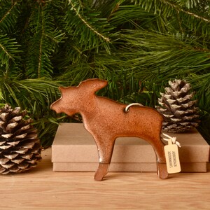 May include: A copper-colored moose ornament with a string for hanging. The ornament is in front of a backdrop of pine needles and two pine cones. A small tag reads "ADIRONDACK ADORNMENTS."