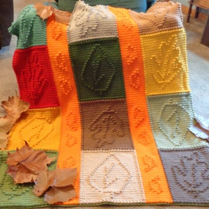 May include: A crocheted blanket with a patchwork design featuring different colored squares with a leaf pattern. The blanket is made with a variety of colors, including green, red, orange, yellow, brown, and beige.