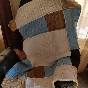Puede incluir: Una manta de bebé de ganchillo con un diseño de patchwork en blanco, marrón, azul y beige. La manta presenta una variedad de texturas y patrones.