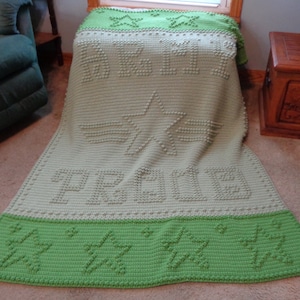 Op de afbeelding: Een gehaakt deken in groen en beige met de woorden "Peace" en "Love" in reliëf. Het deken is versierd met sterren en bloemen.