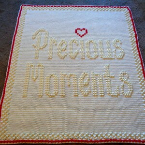 May include: A white crocheted baby blanket with a red border. The blanket has the words "Precious Moments" stitched in yellow yarn with a small red heart above the words.