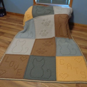 May include: A crocheted blanket with a patchwork design featuring cats, paw prints, and the word "LOVE" in brown, gray, and yellow yarn.