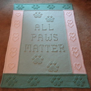 Puede incluir: Una manta de bebé de crochet en tonos verdes y rosas con el texto "All Paws Matter" en el centro. La manta está decorada con huellas de patas y corazones.
